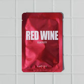 A red-colored face mask pack labeled 'Lapcos Red Wine Elasticity' attached to a white tiled wall.